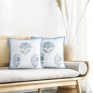 May include: Two square throw pillows with a white background and a blue floral design. The pillows have a blue border and are placed on a light-colored wooden bench. The pillows are decorated with a floral pattern.