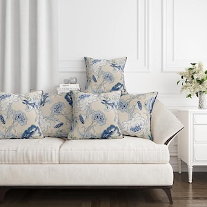 May include: Five decorative throw pillows with a floral pattern in shades of blue and white on a beige background. The pillows are arranged on a white sofa. The room has white walls and a white curtain.