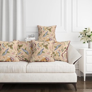 May include: A collection of decorative throw pillows with a floral and peacock design. The pillows feature a beige background with colorful flowers, green leaves, and blue peacocks. The pillows are arranged on a white sofa.