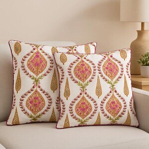 May include: Two decorative throw pillows with a cream background and a repeating pattern of pink, gold, and green floral designs. The pillows have a dark pink border and are placed on a cream-colored sofa.