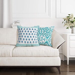 May include: Two decorative throw pillows on a white sofa. One pillow features a blue floral pattern on a white background with a teal border. The other pillow has a teal and white geometric design. The sofa has a dark wood base.