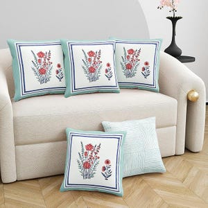 May include: A collection of decorative throw pillows with floral designs. The pillows feature a white background with a red and blue floral pattern, framed by a blue border and a light green edge. The pillows are arranged on a beige sofa.