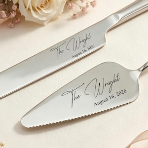 Puede incluir: Un juego de cuchillo y servidor de pastel plateado. El cuchillo tiene una hoja larga y curva, y el servidor tiene una hoja triangular con borde dentado. Ambos están grabados con "The Wright" y "August 16, 2026". El juego se exhibe con flores y cinta.