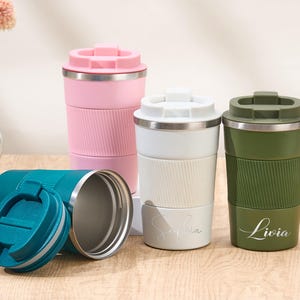 May include: Four stainless steel travel tumblers with lids, featuring a textured middle band. One is teal and lying on its side with the lid detached, while the others are pink, white, and olive green, standing upright with names printed on them.