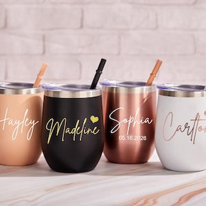 以下が含まれることがあります： ピーチ、黒、ローズゴールド、白の4つのステムレスワインタンブラー。それぞれに筆記体で名前が書かれています。ピーチのタンブラーには「Hayley」、黒には金のハート付きで「Madeline」、ローズゴールドには「Sophia」と日付、白には「Carlton」と書かれています。