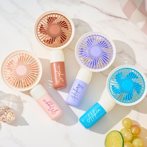 May include: Four handheld portable fans with circular heads and cylindrical handles. The fans feature blades in terracotta, light pink, lavender, and bright blue. Each handle is personalized with white or black script text reading Sophia, Maggie, Hilary, and Alethea.