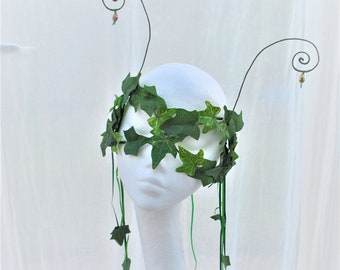 May include: A green ivy leaf mask with two wire tendrils that curl at the top. The mask is on a white mannequin head.