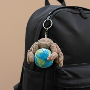 Può includere: Un portachiavi all'uncinetto con una creatura marrone a forma di ragno che tiene in mano un globo blu e verde. Il portachiavi è attaccato a uno zaino nero con una cerniera argentata. Il ragno ha macchie verdi.