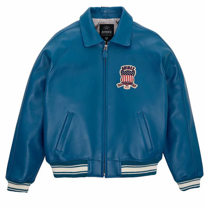 Op de afbeelding: Een felblauwe leren jas in bomberstijl. Het heeft een ritssluiting, een kraag en een borstpatch met een merklogo en een patriottisch ontwerp. De manchetten en zoom zijn afgewerkt met wit en blauwe strepen.
