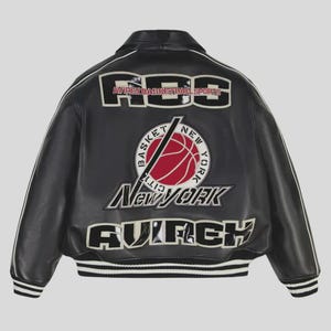 Puede incluir: Chaqueta de cuero negra con detalles blancos y dobladillo acanalado. Presenta el texto "RGS AVIREX BASKETBALL SPORTS" en los hombros y un gr&aacute;fico de baloncesto con el texto "New York". Una chaqueta cl&aacute;sica de tem&aacute;tica deportiva.