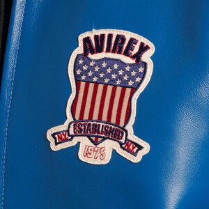 Op de afbeelding: Een close-up van een blauwe leren jas met een patch. De patch toont het woord "AVIREX" boven een schildontwerp met rode en witte strepen en witte sterren. De patch bevat ook de tekst "ESTABLISHED NY 1975 NY".