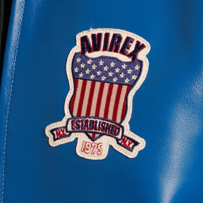 Op de afbeelding: Een close-up van een blauwe leren jas met een patch. De patch toont het woord "AVIREX" boven een schildontwerp met rode en witte strepen en witte sterren. De patch bevat ook de tekst "ESTABLISHED NY 1975 NY".