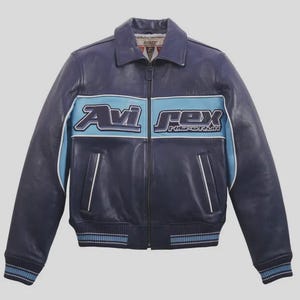 K&ouml;nnte beinhalten: Marineblaue Lederjacke mit hellblauem Streifen auf der Brust, mit dem Text "Avi Rex" in Wei&szlig;. Die Jacke hat einen Kragen, einen Rei&szlig;verschluss und gerippte B&uuml;ndchen und Saum. Klassischer Bomber-Stil.