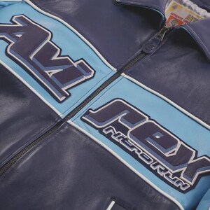 K&ouml;nnte beinhalten: Marineblaue Lederjacke mit hellblauen Akzenten und dem Schriftzug "AVEX NITRO RUN" in einem stilisierten Schriftzug. Die Jacke hat einen Rei&szlig;verschluss und wei&szlig;e Paspeln.