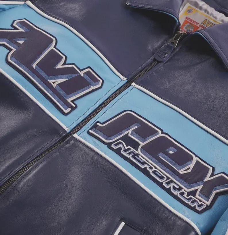 K&ouml;nnte beinhalten: Marineblaue Lederjacke mit hellblauen Akzenten und dem Schriftzug "AVEX NITRO RUN" in einem stilisierten Schriftzug. Die Jacke hat einen Rei&szlig;verschluss und wei&szlig;e Paspeln.