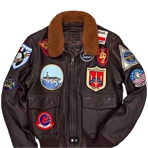 May include: A brown leather bomber jacket with a shearling collar. The jacket features multiple embroidered patches, including military insignia and text. It has two front pockets and a zipper closure.