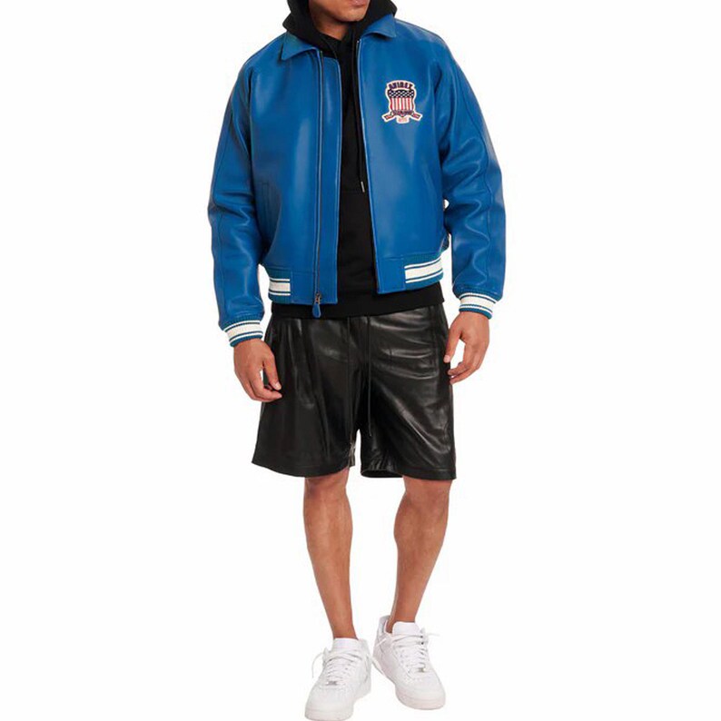 Op de afbeelding: Een felblauwe leren jas met wit en blauw gestreepte kraag en manchetten. De jas heeft een embleem op de borst. De persoon draagt een zwarte korte broek en witte sneakers.