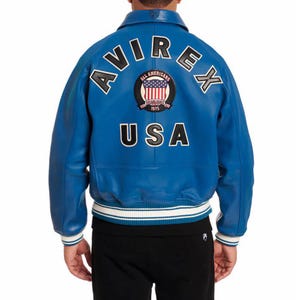 Op de afbeelding: Een felblauwe leren jas in bomberstijl. De achterkant toont het woord "AVIREX" in witte letters, een cirkelvormige patch met de Amerikaanse vlag en het woord "USA". De jas heeft een geribbelde kraag, manchetten en zoom met witte en blauwe strepen.