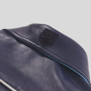 K&ouml;nnte beinhalten: Nahaufnahme einer marineblauen Ledertasche mit einem kleinen, dunklen Emblemaufn&auml;her. Die Tasche hat eine hellblaue Paspel am Rand und eine strukturierte Oberfl&auml;che. Die Tasche scheint aus hochwertigem Leder gefertigt zu sein.