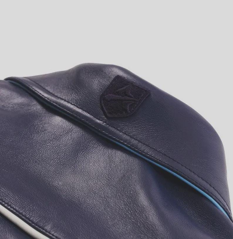 K&ouml;nnte beinhalten: Nahaufnahme einer marineblauen Ledertasche mit einem kleinen, dunklen Emblemaufn&auml;her. Die Tasche hat eine hellblaue Paspel am Rand und eine strukturierte Oberfl&auml;che. Die Tasche scheint aus hochwertigem Leder gefertigt zu sein.