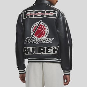 Puede incluir: Chaqueta de cuero negra con ribetes blancos y el texto "AVIREX BASKETBALL SPORTS" en los hombros. Un gr&aacute;fico de baloncesto con "New York" est&aacute; centrado en la espalda. La chaqueta tiene dobladillo y pu&ntilde;os acanalados.