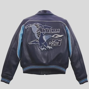 K&ouml;nnte beinhalten: Marineblaue Lederjacke mit einem gro&szlig;en Adleraufn&auml;her auf dem R&uuml;cken. Der Aufn&auml;her zeigt den Text "Avirex", "NY75" und ein Adlerdesign in Blau- und Wei&szlig;t&ouml;nen. Hellblaue Streifen verlaufen die &Auml;rmel entlang.
