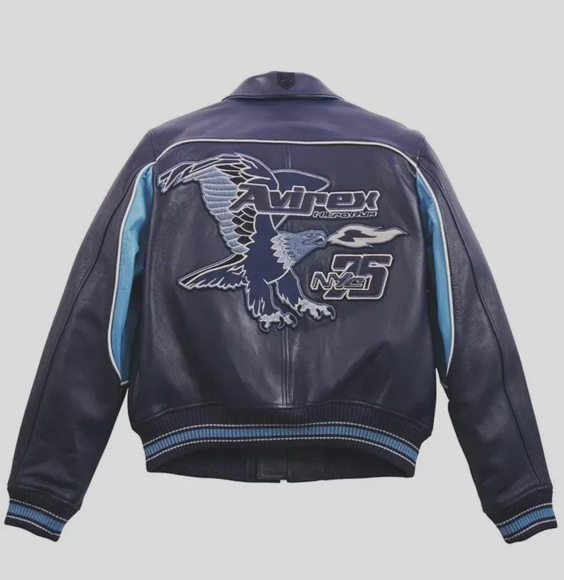 K&ouml;nnte beinhalten: Marineblaue Lederjacke mit einem gro&szlig;en Adleraufn&auml;her auf dem R&uuml;cken. Der Aufn&auml;her zeigt den Text "Avirex", "NY75" und ein Adlerdesign in Blau- und Wei&szlig;t&ouml;nen. Hellblaue Streifen verlaufen die &Auml;rmel entlang.