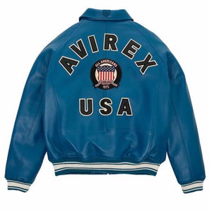 Op de afbeelding: Een blauwe leren jas met het woord "AVIREX" in witte letters over de schouders. Daaronder een cirkelvormige patch met de Amerikaanse vlag en de woorden "ALL AMERICANS ESTABLISHED 1975". Het woord "USA" staat eronder.