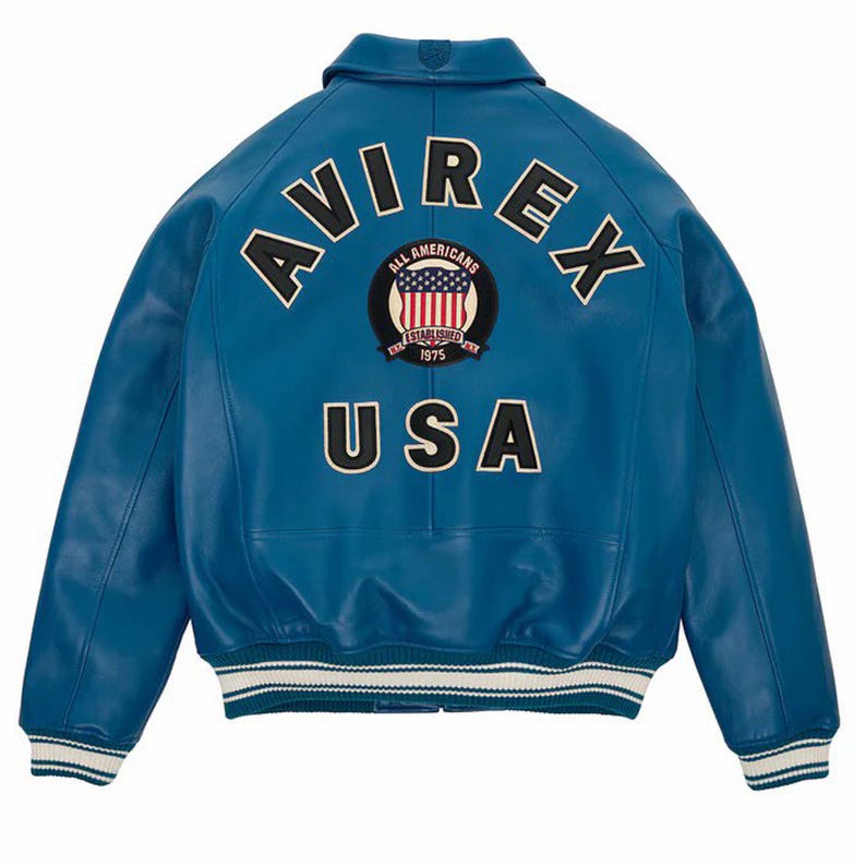 Op de afbeelding: Een blauwe leren jas met het woord "AVIREX" in witte letters over de schouders. Daaronder een cirkelvormige patch met de Amerikaanse vlag en de woorden "ALL AMERICANS ESTABLISHED 1975". Het woord "USA" staat eronder.