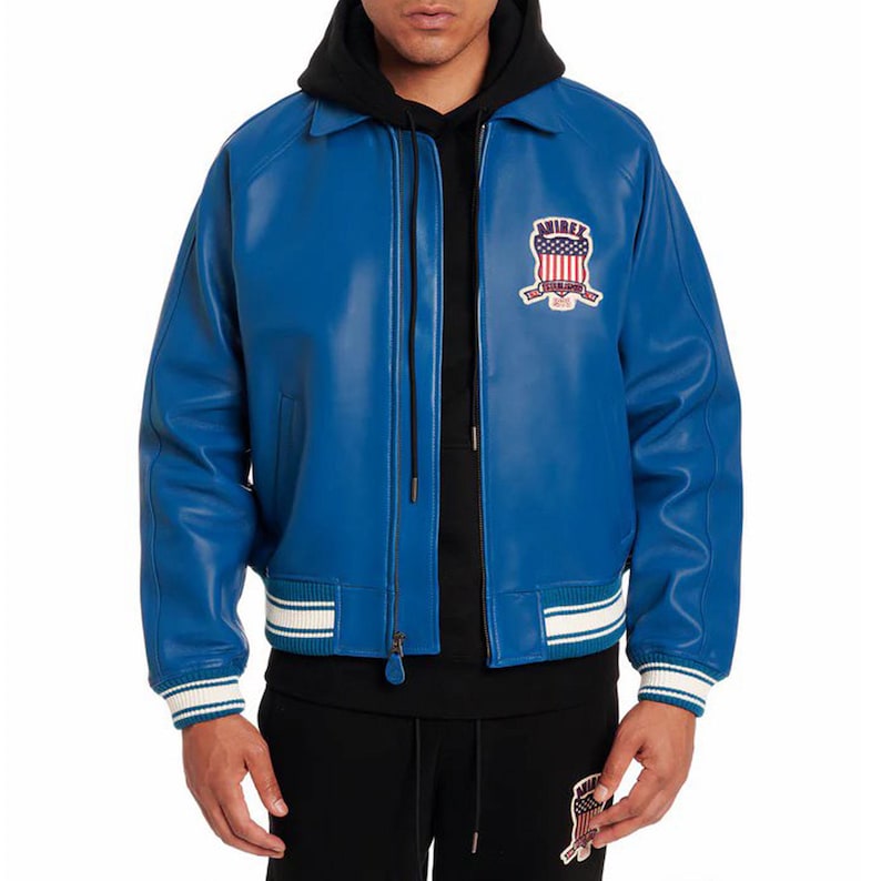 Op de afbeelding: Felblauw leren jack met ritssluiting, wit en blauw gestreepte kraag, manchetten en zoom. Het jack heeft een borstpatch met de tekst "AVIREX" en een patriottisch ontwerp.