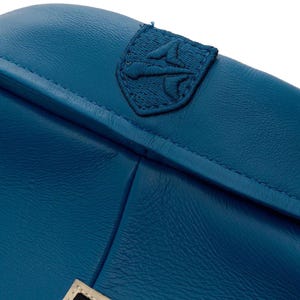 Op de afbeelding: Close-up van een tealblauwe leren tas met een gestikt embleem. De tas heeft een getextureerd oppervlak en zichtbare stiksels langs de naden. Het embleem is een donkerdere tint blauw en is geborduurd.