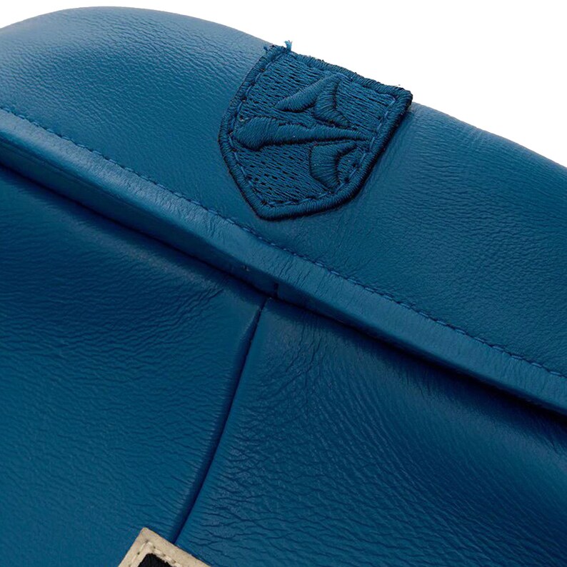 Op de afbeelding: Close-up van een tealblauwe leren tas met een gestikt embleem. De tas heeft een getextureerd oppervlak en zichtbare stiksels langs de naden. Het embleem is een donkerdere tint blauw en is geborduurd.