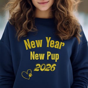 Op de afbeelding: Marineblauw sweatshirt met de tekst "New Year New Pup 2026" in het geel. Een geel hart met een pootafdruk is ook afgebeeld. Het sweatshirt heeft een casual stijl.