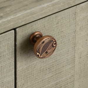 May include: Close-up of a bronze cabinet knob with an intricate, patterned design. The knob is round with a detailed surface, mounted on a light gray wooden cabinet door. The cabinet door has a textured, wood-grain appearance.