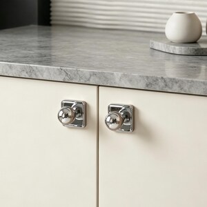 May include: Two polished chrome cabinet knobs with a spherical handle and a square backplate, installed on light cream-coloured cabinet doors. The knobs feature a reflective metallic finish and a simple, clean design for furniture hardware.