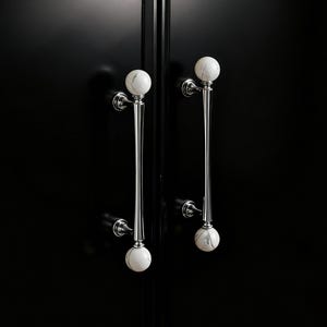 May include: Two cabinet door handles with a polished chrome finish and white marble sphere accents. The handles are long and slender, with a decorative design. The cabinet doors are black.