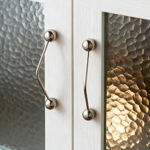 Peut inclure: Gros plan sur une armoire blanche à deux portes avec des panneaux de verre texturés. Chaque porte est équipée d'une poignée argentée incurvée avec une extrémité sphérique. L'armoire semble être en bois.