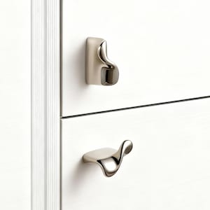 May include: Close-up of a white cabinet with two silver-toned drawer pulls. The top pull is a small, curved handle, while the bottom pull has a more elaborate, flowing design. The cabinet has a vertical, grooved trim on the left side.