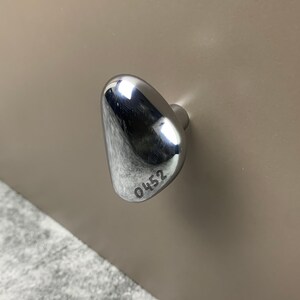 May include: A polished chrome cabinet knob with an irregular, organic pebble shape. The surface is highly reflective and features the engraved number 0452 near the bottom edge. The knob is mounted on a smooth, light beige flat surface.