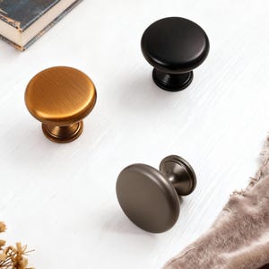 Peut inclure: Trois boutons de meubles de différentes finitions : bronze, noir et gris. Les boutons ont un dessus rond et une base décorative. Les boutons sont sur une surface blanche avec un livre et un tissu.
