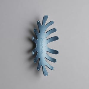 May include: A blue hair claw clip with a curved, organic, leaf-like shape featuring multiple rounded prongs. The surface has a smooth, matte finish and displays the text 3NordicVest in a vertical orientation along the central spine of the clip.