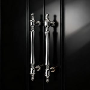 May include: Two polished chrome door handles with a decorative design are mounted on a black door. The handles have a long, slender shape with ornate detailing at the top and bottom. The door's surface is glossy.