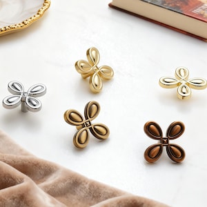 May include: Five cabinet knobs in silver, gold, bronze, and copper finishes. Each knob has a four-petal design with a square centre. These decorative knobs are suitable for furniture or home decor projects.