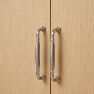 Könnte beinhalten: Zwei Schrankgriffe aus gebürstetem Nickel mit geschwungenem Design sind an einer hellen Holzschranktür montiert. Die Griffe haben eine zylindrische Mitte und gebogene Enden und bieten eine moderne und funktionale Ästhetik.