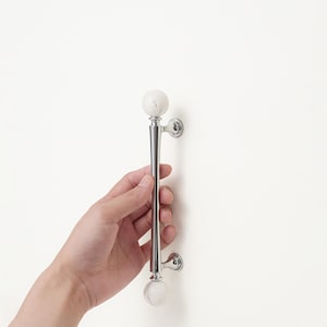 May include: A polished chrome cabinet pull with white marble-like spherical ends. The handle is held against a white surface. The pull is approximately 25 cm long.