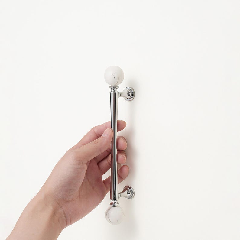 May include: A polished chrome cabinet pull with white marble-like spherical ends. The handle is held against a white surface. The pull is approximately 25 cm long.