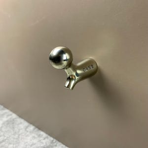 Puede incluir: Un gancho de pared de metal dorado pulido con una cabeza esférica y un cuerpo curvo. El lateral del gancho tiene grabado el número 0452. El gancho está montado sobre una superficie lisa de color marrón claro que ocupa el encuadre.