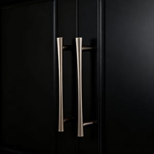 Könnte beinhalten: Nahaufnahme von zwei Türgriffen aus gebürstetem Nickel an einem schwarzen Schrank. Die Griffe sind vertikal, mit einem sich verjüngenden Design, und sind auf einer dunklen, festen Oberfläche montiert. Die Hardware hat eine moderne, minimalistische Ästhetik.