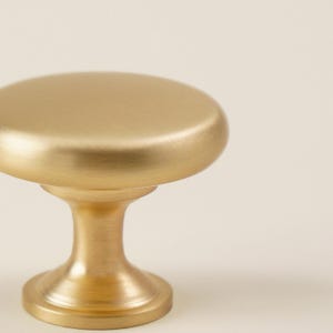 Pu&ograve; includere: Primo piano di una manopola per mobili color oro. La manopola ha una parte superiore arrotondata e una base a piedistallo. La finitura &egrave; opaca e l'hardware &egrave; probabilmente in metallo. Lo sfondo &egrave; di un colore bianco sporco.