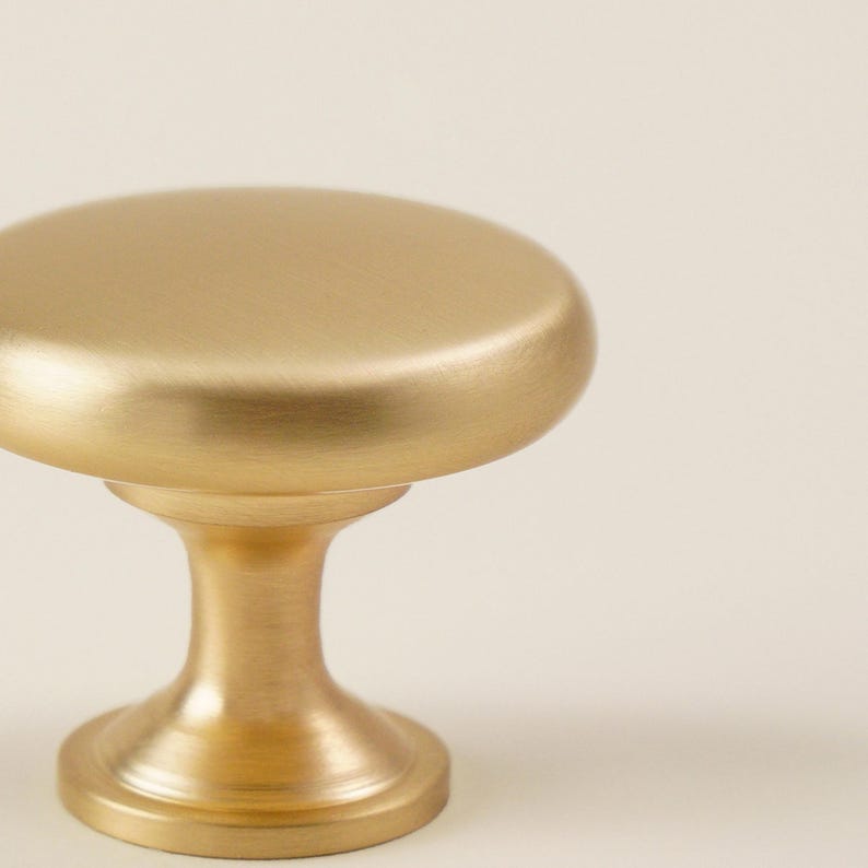 Pu&ograve; includere: Primo piano di una manopola per mobili color oro. La manopola ha una parte superiore arrotondata e una base a piedistallo. La finitura &egrave; opaca e l'hardware &egrave; probabilmente in metallo. Lo sfondo &egrave; di un colore bianco sporco.